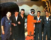 טייס פנטום מטייסת 119 לובש חליפת לחץ לטיסות בגובה מעל ל-70,000 רגל בצבע כתום, לידו עומדים אנשי צוות קרקע בסרבלי עבודה כחולים.